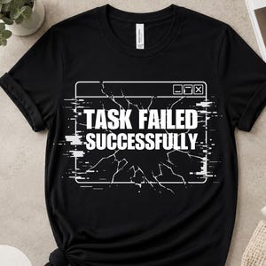 Puede incluir: Camiseta negra con el texto blanco "TASK FAILED SUCCESSFULLY" dentro de un gráfico de ventana de computadora rota. La camiseta está doblada en la parte inferior y tiene cuello redondo.