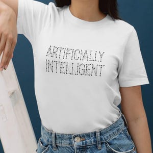 Puede incluir: Camiseta blanca con la frase "ARTIFICIALLY INTELLIGENT" en negro, diseñada con puntos conectados. La camiseta se combina con vaqueros azules. El estilo general es casual y moderno.