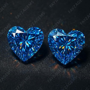 Könnte beinhalten: Zwei herzförmige, brillantgeschliffene Edelsteine. Die Edelsteine sind leuchtend blau, mit Facetten, die das Licht einfangen, um einen funkelnden Effekt zu erzeugen. Der Hintergrund ist dunkel und betont die Farbe und Form der Edelsteine. Der Text "FAYZAGEMS" ist sichtbar.