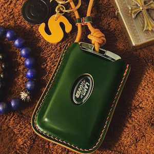 May include: A dark green leather Land Rover key fob cover with orange stitching. The fob is on a brown surface with a leather keychain, a gold-coloured lighter, and a beaded bracelet.