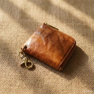 Può includere: Un piccolo portafoglio quadrato in pelle marrone con chiusura a zip e un fermaglio color ottone. Il portafoglio ha una superficie strutturata ed è appoggiato su uno sfondo di juta.