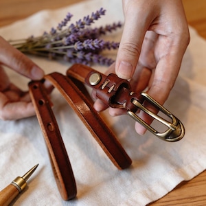 Puede incluir: Un cinturón de cuero marrón con hebilla de latón es sostenido. El cinturón está parcialmente desabrochado, mostrando los detalles del cuero y las costuras. También se ve una ramita de lavanda y una herramienta de trabajo del cuero.
