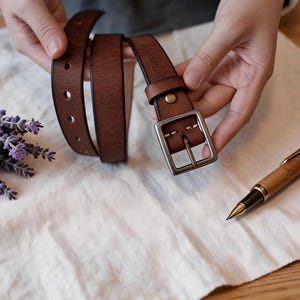 Op de afbeelding: Een bruine leren riem met een vierkante metalen gesp met een zilverkleurige afwerking. De riem heeft een glad, gestructureerd oppervlak met zichtbare stiksels bij de gesp en cirkelvormige gaten die in de lengte van het leer zijn geponst.