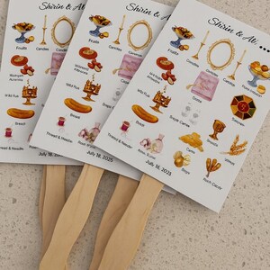 May include: Three white paper fans with watercolor illustrations and text. Each fan features images of fruits, candles, and other decorative elements. The fans have wooden handles and the text "Shirin & Ali" is visible.