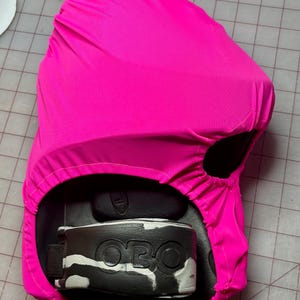 Puede incluir: Una funda protectora rosa intenso para un equipo deportivo. La funda está colocada sobre un objeto en blanco y negro con las letras "OBO" visibles. El equipo parece ser un equipo de protección.