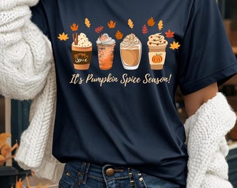 Może przedstawiać: Granatowa koszulka z nadrukiem czterech pumpkin spice latte z bitą śmietaną i jesiennymi liśćmi. Tekst "It's Pumpkin Spice Season!" jest wydrukowany pod grafiką latte. Koszulka jest połączona z niebieskimi dżinsami.
