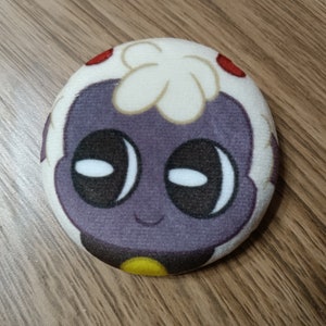 May include: A plush toy shaped like a round character with a purple face, white eyes, and a white fluffy head. The character has a small yellow detail on its chest.