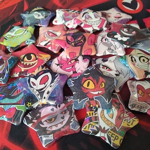 May include: A collection of 16 star-shaped pins featuring various characters from the animated series "Hazbin Hotel". The pins are colorful and feature a variety of designs, including characters with horns, wings, and other unique features.