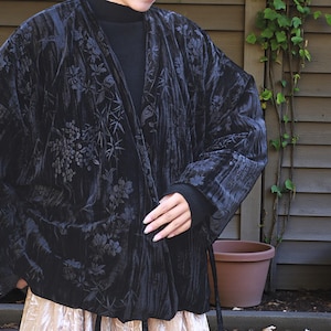 Könnte beinhalten: Schwarze Samt-Kimono-Jacke mit floralem Muster. Die Jacke hat lange Ärmel und einen Bindverschluss. Das Model trägt einen schwarzen Rollkragenpullover und einen beigen Rock. Der Hintergrund umfasst eine Holzwand und Blumentöpfe.