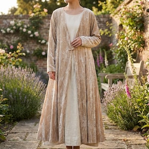 May include: A long-sleeved, floor-length beige velvet duster coat featuring a subtle white floral pattern and a single front toggle closure, worn over a simple, solid cream-coloured floor-length dress with a round neckline and long hem.