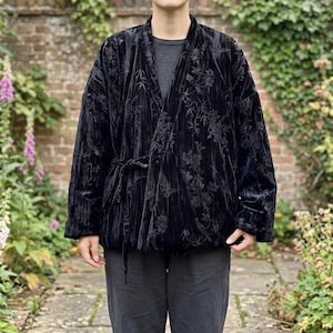 以下が含まれることがあります： 黒いベルベット素材のラップジャケット。同系色の花柄と植物柄が施され、長袖でサイドに結び紐が付いています。ダークグレーのシャツとダークグレーのパンツの上に重ね着されています。