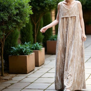 May include: A beige, floor-length jumpsuit with a V-neck and spaghetti straps. The fabric has a subtle floral pattern and a soft, velvety texture. The jumpsuit is loose-fitting and appears comfortable, suitable for casual wear.