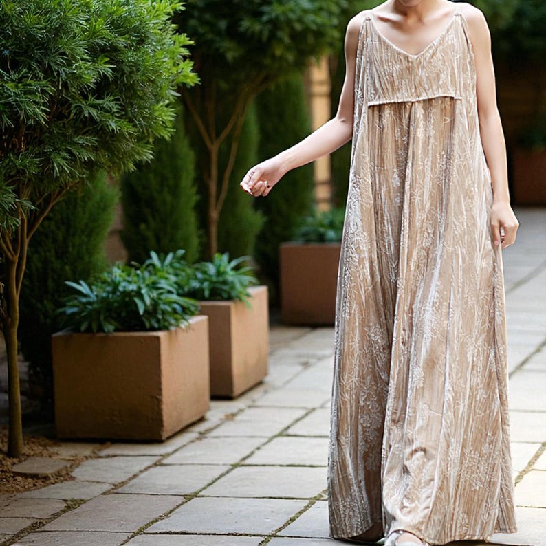May include: A beige, floor-length jumpsuit with a V-neck and spaghetti straps. The fabric has a subtle floral pattern and a soft, velvety texture. The jumpsuit is loose-fitting and appears comfortable, suitable for casual wear.