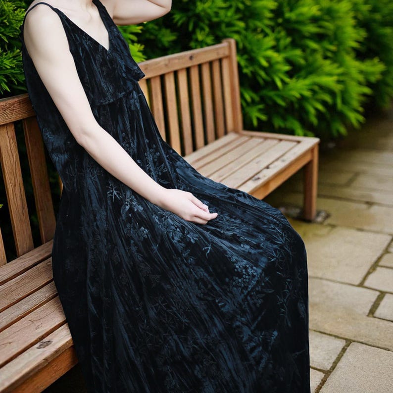 May include: A black velvet maxi dress with a V-neck and spaghetti straps. The dress features a floral pattern and is worn while seated on a wooden bench. The background includes green foliage and a stone walkway.