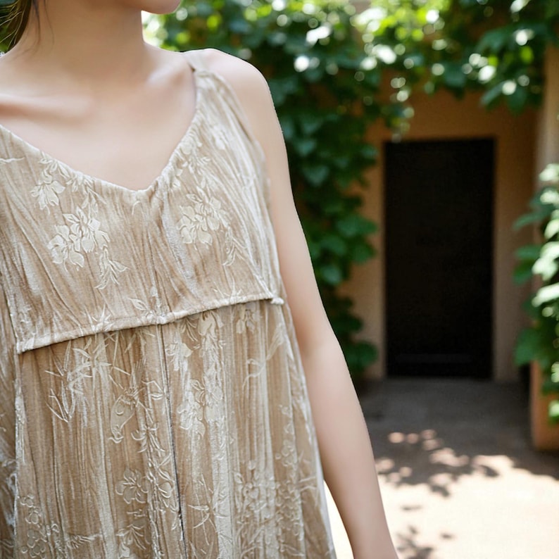 May include: A beige, sleeveless dress with a V-neck and floral pattern. The dress has a textured appearance and a relaxed fit. The background features greenery and a dark doorway, suggesting an outdoor setting. The dress appears to be made of a soft, flowing fabric.