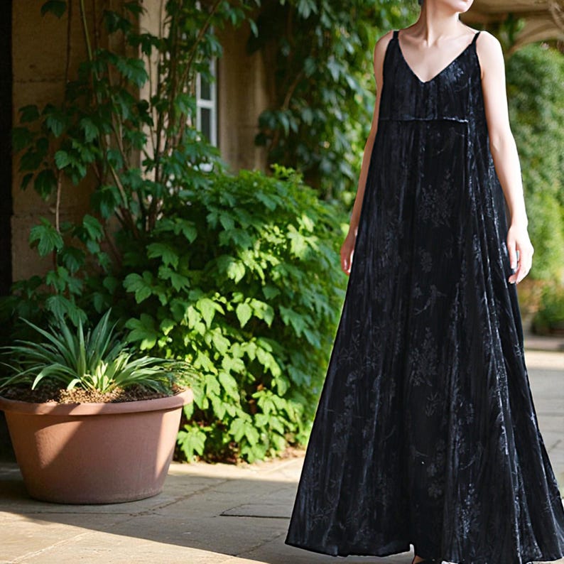 May include: A long, black velvet dress with a V-neck and spaghetti straps. The dress has a floral pattern and a flowing silhouette. The model is standing outside in front of lush greenery.
