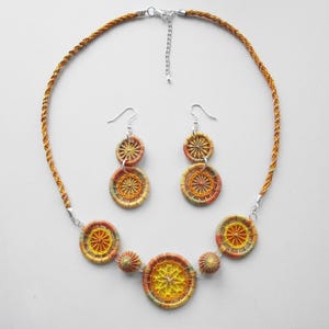 May include: A handcrafted jewelry set featuring a necklace and matching earrings. The necklace and earrings have a circular design with yellow, orange, and brown woven details. The necklace has a braided cord and a silver-toned clasp.