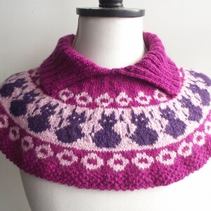 May include: A knitted cowl in shades of fuchsia, purple, and white, displayed on a mannequin. The cowl features a repeating pattern of cat silhouettes and floral designs. The collar is a solid fuchsia color.