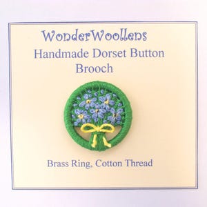Puede incluir: Broche de botón Dorset hecho a mano con un borde verde, con un ramo de flores azules y un lazo amarillo. El texto en la tarjeta dice "WonderWoollens" y "Brass Ring, Cotton Thread."