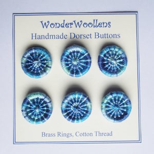 Puede incluir: Seis botones Dorset hechos a mano en tonos azules y verdes, dispuestos sobre una tarjeta crema. Los botones están hechos con aros de latón e hilo de algodón. La tarjeta dice "WonderWoollens Handmade Dorset Buttons."