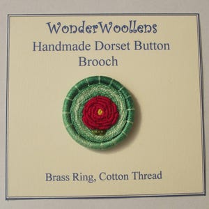 Puede incluir: Broche Dorset Button hecho a mano con un centro de rosa roja y anillos exteriores verdes. Fabricado con un anillo de latón e hilo de algodón. El texto en la tarjeta dice "WonderWoollens".