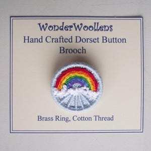 Puede incluir: Broche de botón Dorset hecho a mano con un diseño de arcoíris. El broche está hecho con un anillo de latón e hilo de algodón. El texto en la tarjeta dice "WonderWoollens Hand Crafted Dorset Button Brooch".