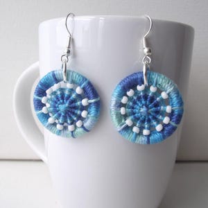 Puede incluir: Un par de pendientes tejidos a mano en azul y blanco. Cada pendiente circular presenta un diseño central con cuentas blancas y un patrón tejido en tonos azules. Los pendientes cuelgan de ganchos plateados.