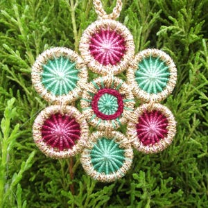 May include: A handmade pendant necklace featuring a floral design. The pendant has a gold-colored border with alternating maroon and teal circular elements. The center of each circle has a starburst pattern. The necklace chain is gold-colored.