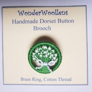 Puede incluir: Broche Dorset Button hecho a mano con un diseño verde y blanco. El broche presenta un anillo bordado verde con un diseño floral blanco y un lazo blanco. El texto en la tarjeta dice "WonderWoollens" y "Brass Ring, Cotton Thread."