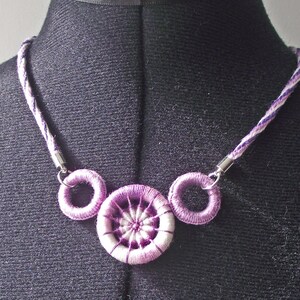 Puede incluir: Collar hecho a mano con un diseño morado y blanco. El collar tiene un cordón trenzado con un patrón morado y blanco. El colgante tiene tres elementos circulares, con un círculo central bordado más grande y dos círculos más pequeños.