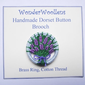 Puede incluir: Broche de botón Dorset hecho a mano con un diseño de ramo de lavanda. El broche circular tiene un borde tejido azul claro y verde. La lavanda es morada y el lazo es morado oscuro. El texto en la tarjeta dice "WonderWoollens".