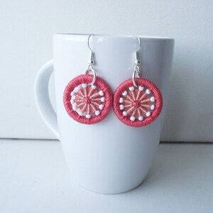 May include: A pair of handcrafted earrings with a circular design. The earrings have a coral-coloured woven outer ring, with a lighter coral and white embroidered pattern in the centre. They hang from silver-coloured hooks.