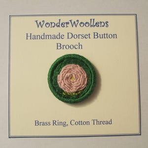 Puede incluir: Broche de botón Dorset hecho a mano con un diseño de rosa rosa en el centro, rodeado por un borde verde. El texto en la tarjeta dice "WonderWoollens Handmade Dorset Button Brooch". Hecho con un anillo de latón e hilo de algodón.