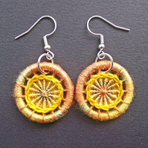 Puede incluir: Un par de pendientes hechos a mano con un diseño circular. Los pendientes presentan un patrón tejido en tonos de amarillo, naranja y verde, con ganchos plateados. Los pendientes se muestran sobre un fondo oscuro.