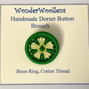Puede incluir: Broche Dorset Button hecho a mano, verde y dorado. El broche circular presenta un anillo exterior verde y un patrón dorado en forma de estrella en el centro. El texto en la tarjeta dice "WonderWoollens Handmade Dorset Button Brooch".