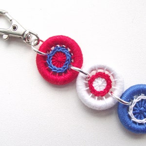 Peut inclure: Un porte-clés fait à la main avec trois ornements circulaires brodés en rouge, blanc et bleu. Les ornements sont reliés par des anneaux argentés et un fermoir. Le design utilise des couleurs patriotiques.