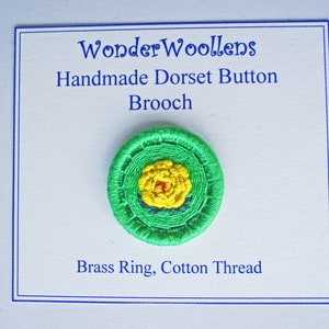 Puede incluir: Broche de botón Dorset hecho a mano con un anillo de latón e hilo de algodón. El broche presenta un diseño floral amarillo en el centro, rodeado de círculos concéntricos de hilo verde. El texto "WonderWoollens" y "Handmade Dorset Button Brooch" son visibles.