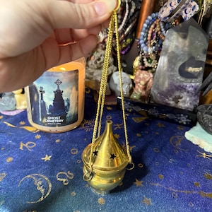 Könnte beinhalten: Ein goldfarbener Räuchergefäß mit konischem Deckel und sternförmigen Aussparungen, der an einer Kette hängt. Der Brenner wird von einer Hand gehalten. Eine Kerze mit dem Text "Ghost Cemetery" befindet sich im Hintergrund.