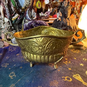 May include: An antique brass bowl with detailed floral and scrollwork. It features two handles and stands on three small feet. The bowl's interior is visible, and it's displayed with other decorative objects.