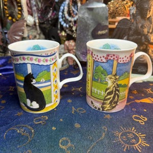 May include: Two white ceramic mugs with cat illustrations. One mug features a black and white cat, the other a tabby cat. Both mugs have a colorful window design and floral accents.