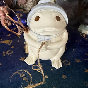Può includere: Un contenitore in ceramica a forma di rana con cappello e papillon azzurro. La rana ha occhi marroni e tiene un paio di forbici decorative. Un pezzo di spago pende dal contenitore.