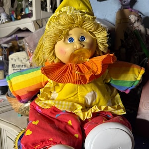 May include: A vintage clown doll with a yellow hat and hair, wearing a yellow shirt with an orange ruffled collar and rainbow sleeves. The doll has a pacifier and red pants with heart patterns. The doll's white shoes are visible.