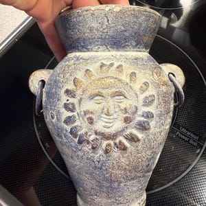 May include: A blue and beige ceramic vase with a sun face design. The vase has two handles and a textured surface. The sun face is in the center of the vase, with a smiling face and sun rays.