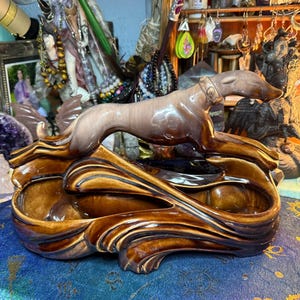 May include: A brown ceramic ashtray with a greyhound dog figurine. The ashtray has a flowing, wave-like design with two recessed areas. The dog is in a running pose, with a collar.