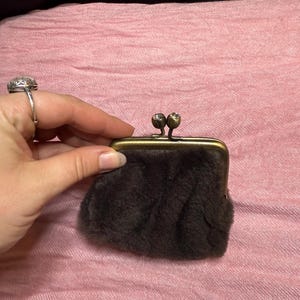 Può includere: Un piccolo portamonete peloso marrone con chiusura in ottone. Il portamonete è tenuto su uno sfondo rosa. La chiusura ha due piccoli elementi decorativi rotondi. Il portamonete è piccolo.