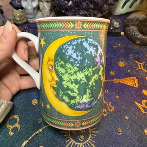 May include: A ceramic mug featuring a crescent moon with a face and a colorful planet design. The mug has a white handle and a green background with stars. The rim is decorated with floral and geometric patterns.