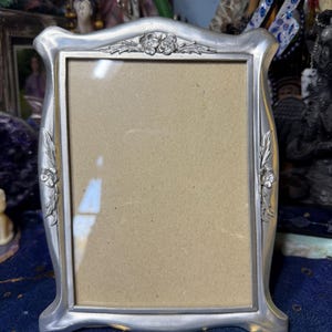 Puede incluir: Un marco de fotos plateado con una abertura rectangular. El marco tiene un diseño floral decorativo en la parte superior y en los lados. El marco es de metal y tiene un aspecto vintage.