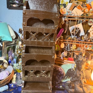 Puede incluir: Un organizador de pared alto de madera marrón oscuro con múltiples estantes y un pequeño cajón. El organizador tiene una parte superior decorativa y presenta un diseño en forma de X en algunos estantes. El diseño general es rústico y vintage.