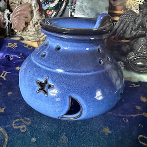 May include: A blue ceramic oil warmer with a dark brown rim and handle. The warmer features a star and crescent moon cutout design, along with several small holes. The oil warmer is a decorative item.
