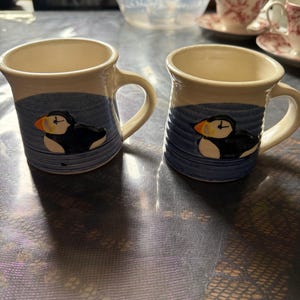 May include: Two ceramic mugs with a cream-colored interior and a blue exterior design featuring a puffin. The mugs have a handle and are set against a dark background. The puffin has a black body, white face, and orange beak.